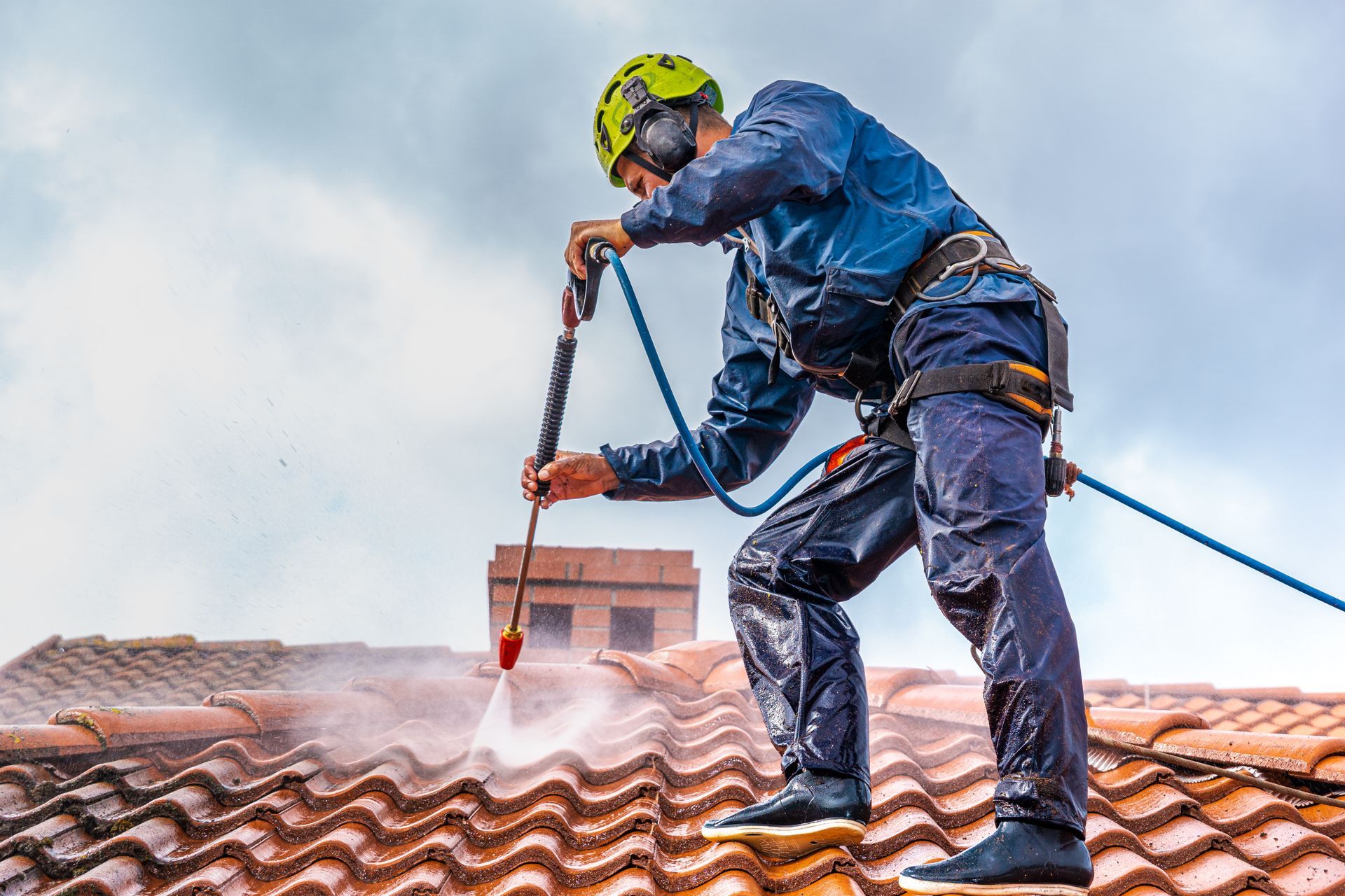 A worker in safety gear pressure-washes a tiled roof while tethered by a rope against a cloudy sky.