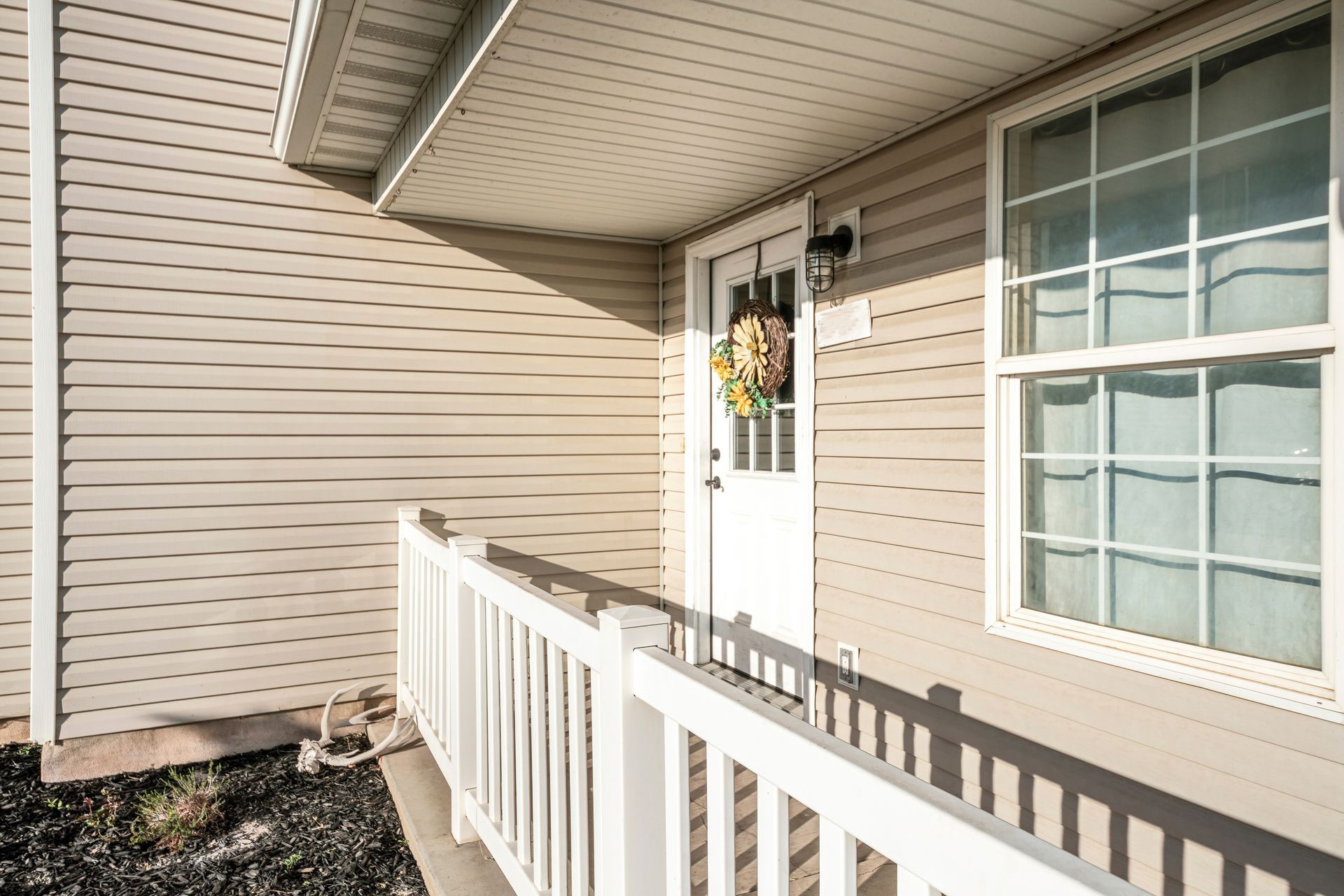 A tan-sided house exterior features a white front door with a wreath, a white railing, and a window with white trim.