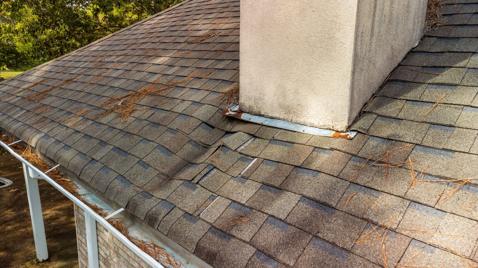 A residential roof with asphalt shingles and a stone chimney featuring damaged, rusted metal flashing.