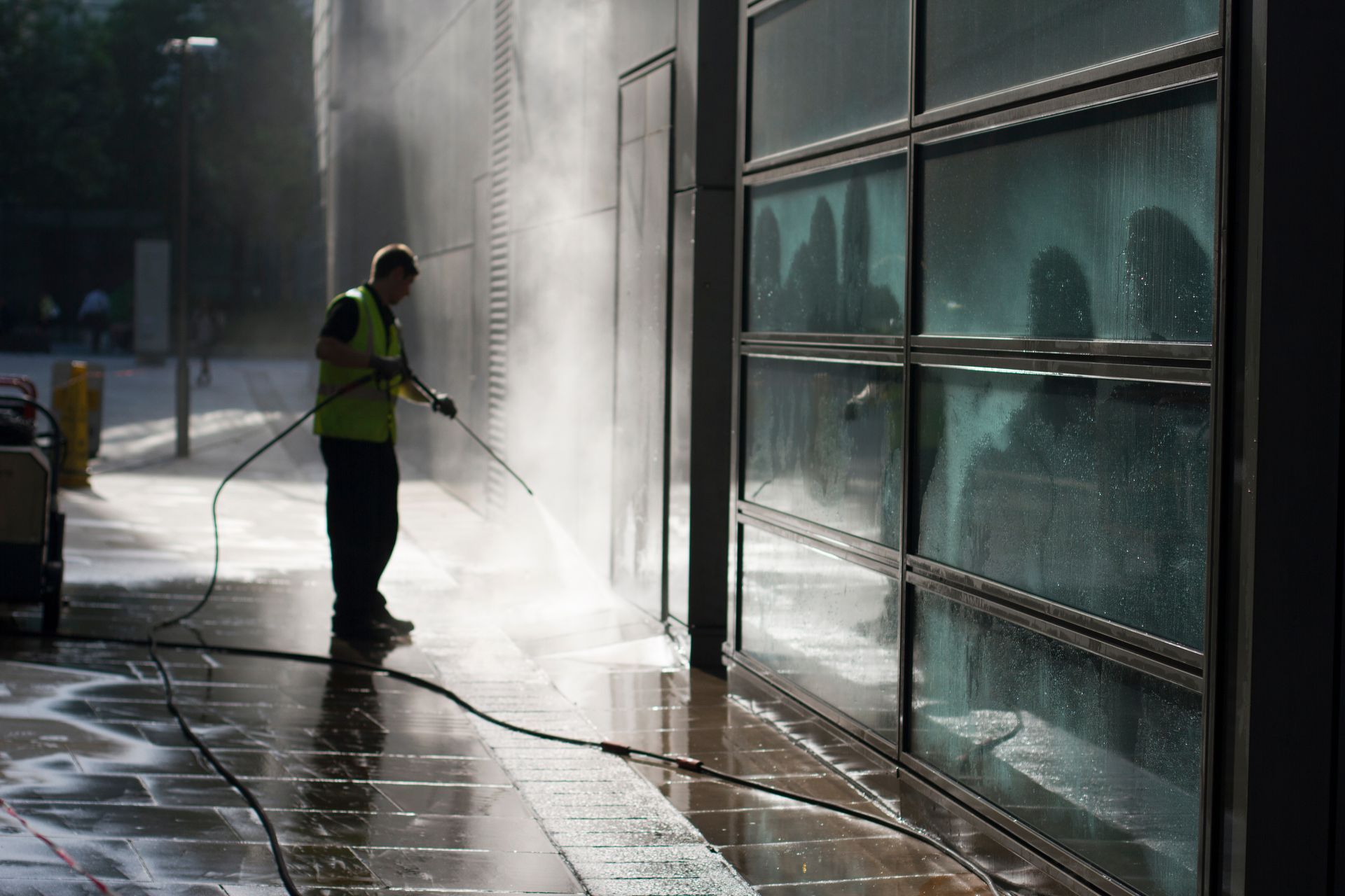 A person in a high-visibility vest uses a pressure washer to clean a building’s exterior glass wall.
