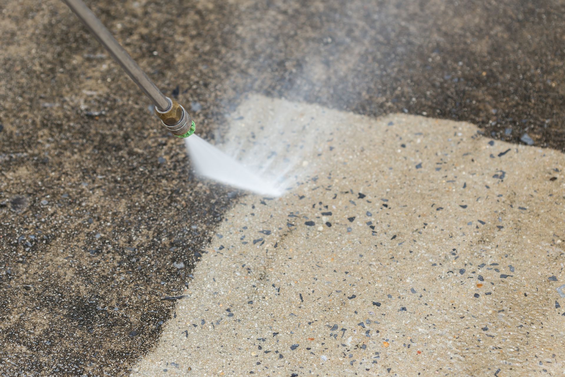 A pressure washer nozzle sprays a stream of water onto a dirty concrete surface, revealing a clean, light-colored patch.