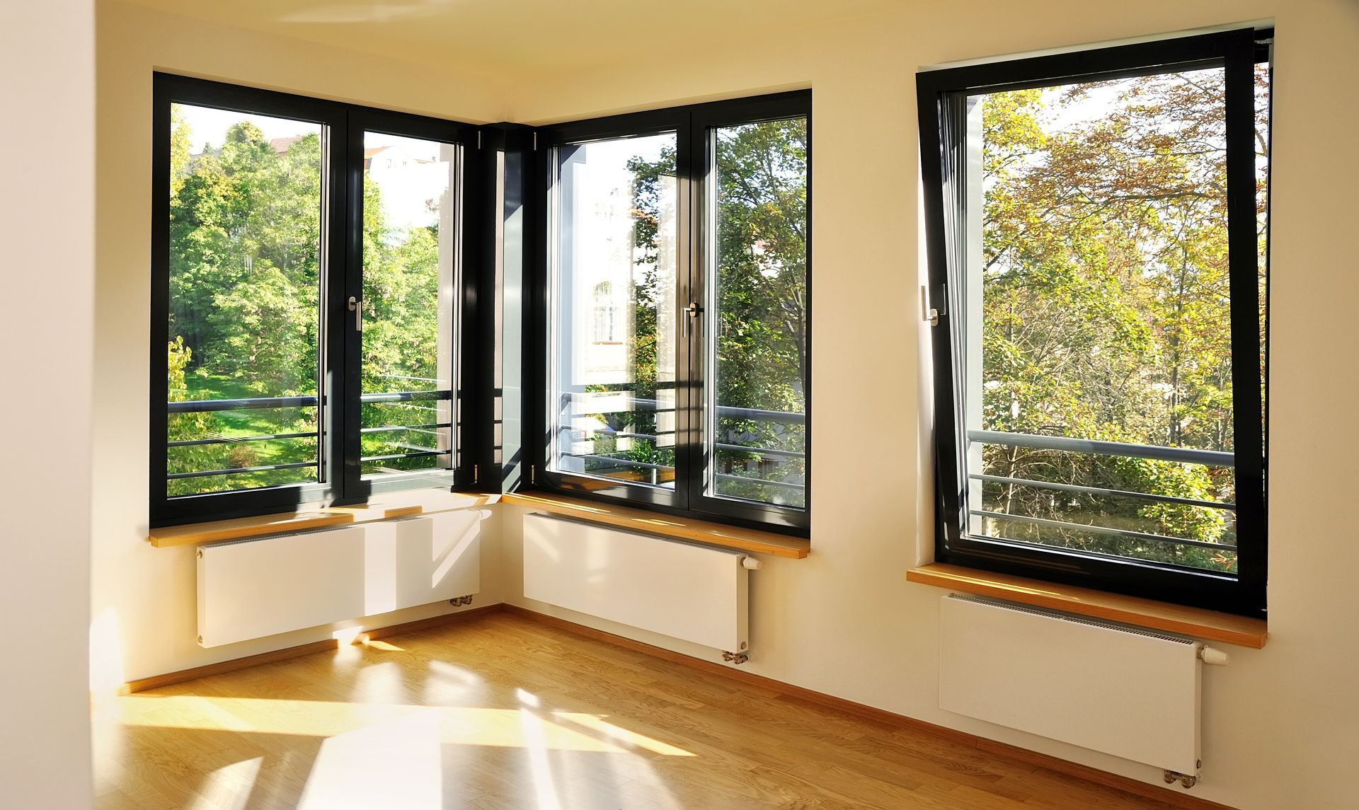 A corner room with wooden floors, white walls, black-framed windows, and radiators placed underneath each window.