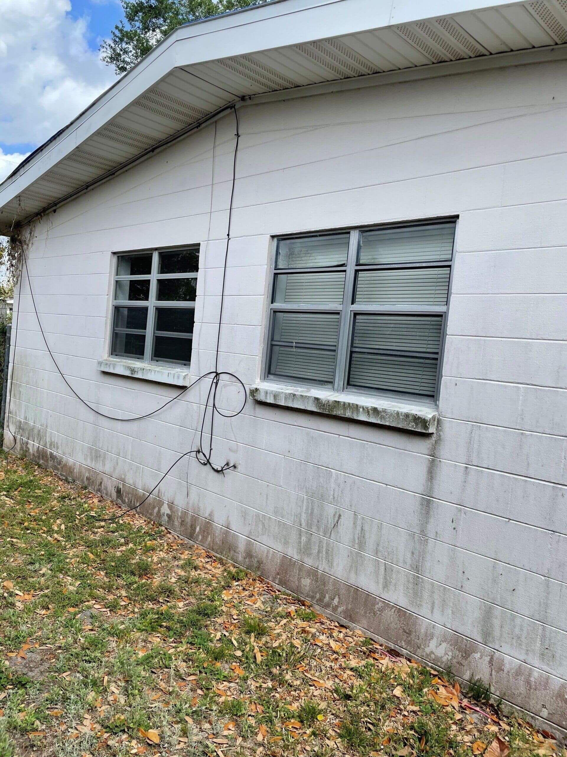 A side view of a white, concrete block exterior wall with two windows and a loose, hanging black cable.