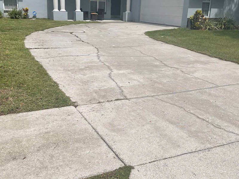 A light gray concrete driveway with visible cracks, flanked by grass, leading to a house entrance.