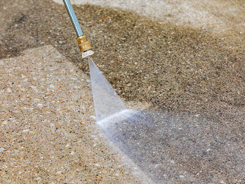 A pressure washer spraying water on a concrete surface, revealing a clean section against a dirty, stained area.