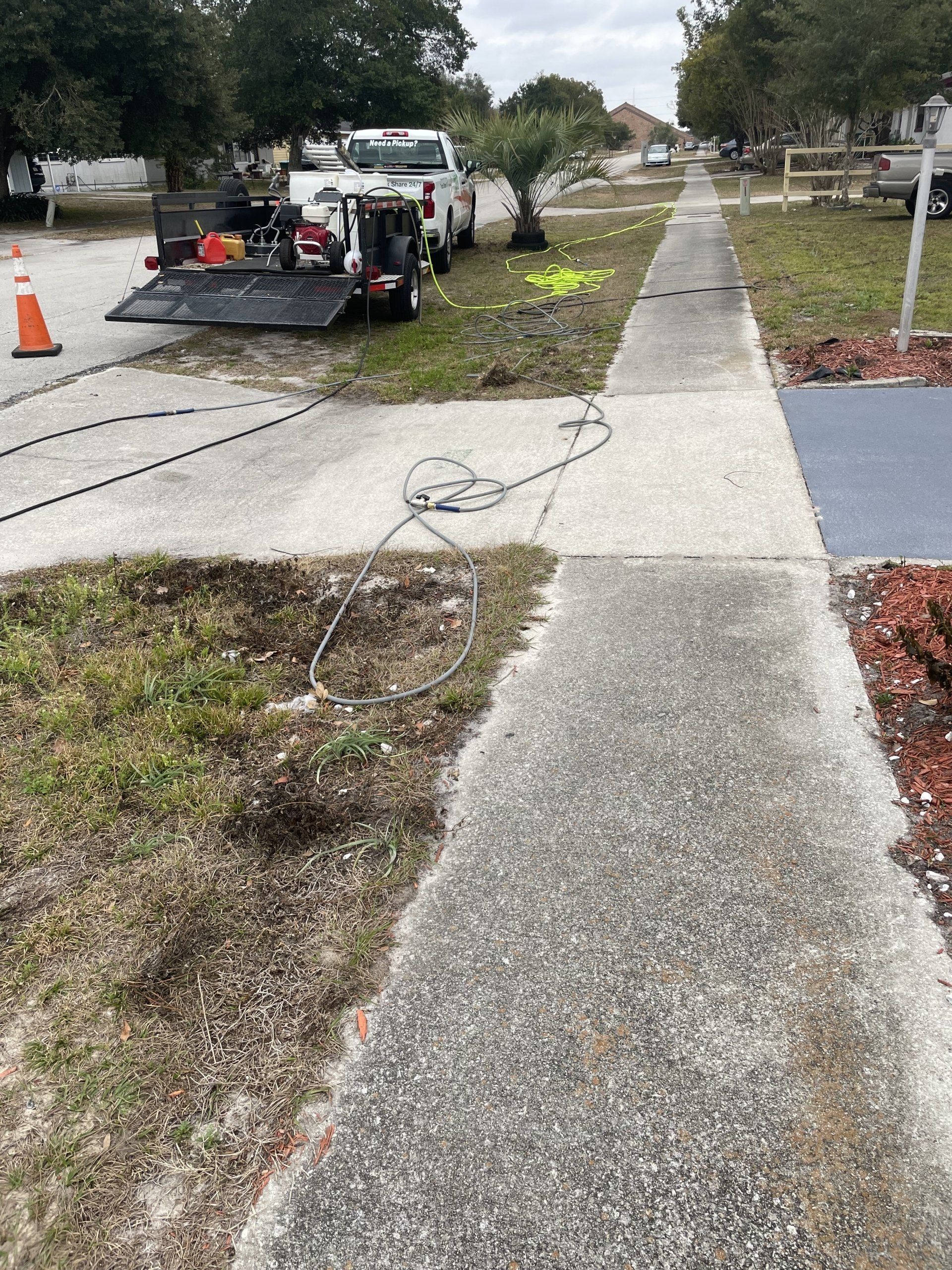 A white pickup truck with a trailer parked on a suburban lawn, with hoses extending across a concrete sidewalk.