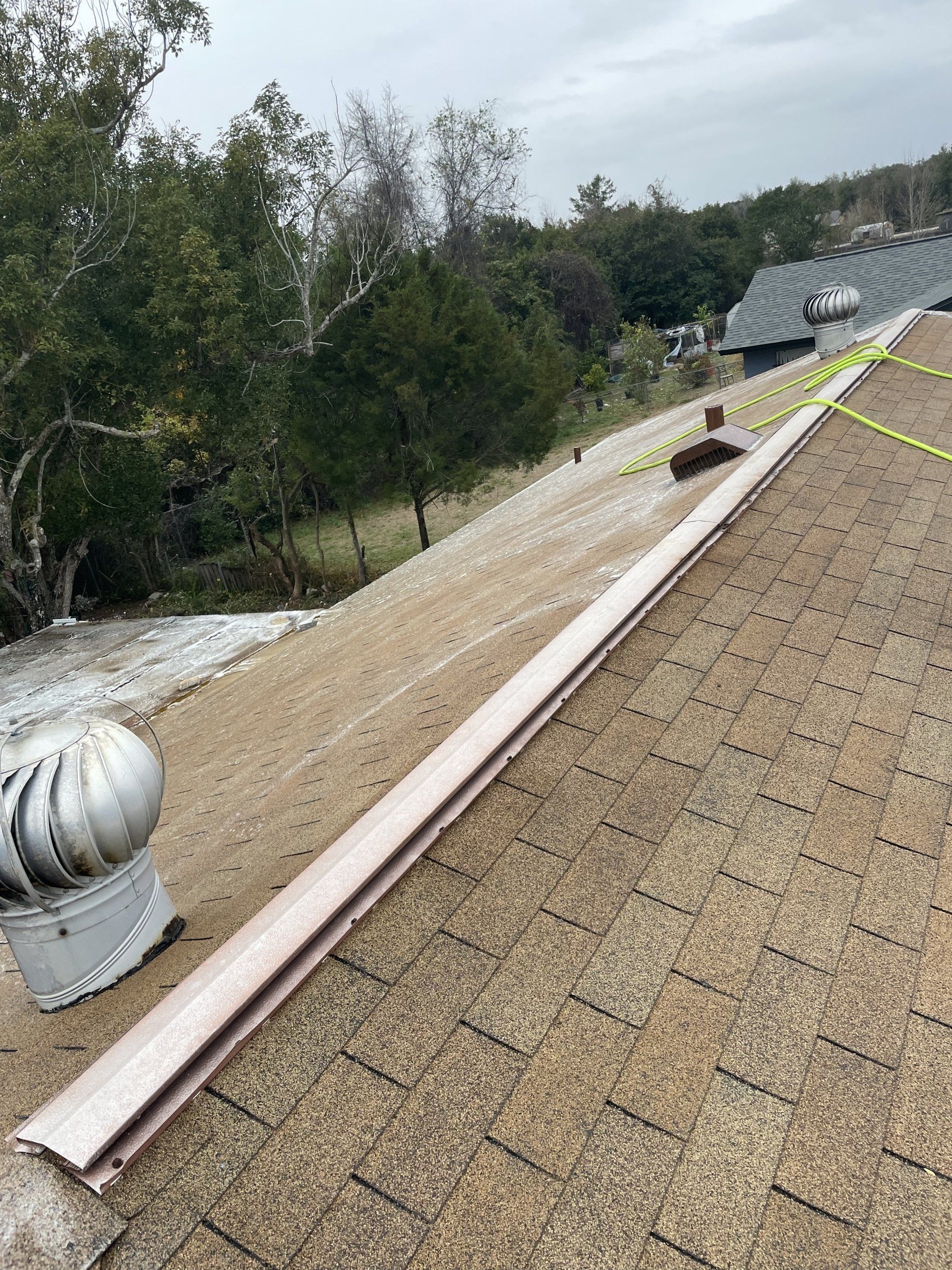 A sloped roof with tan shingles, two metal roof vents, and a long vent ridge strip, set against a background of trees.