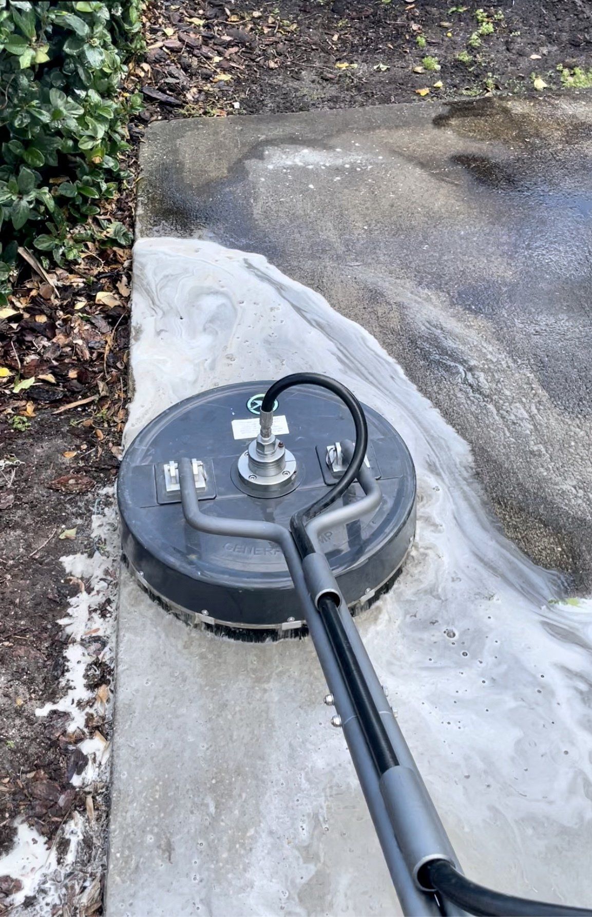 A gray pressure washer surface cleaner cleans a dirty concrete walkway, leaving a path of clean pavement.