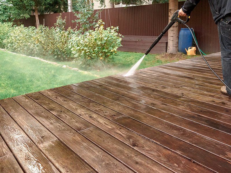 A person uses a pressure washer to clean dark wooden deck planks in a grassy backyard.