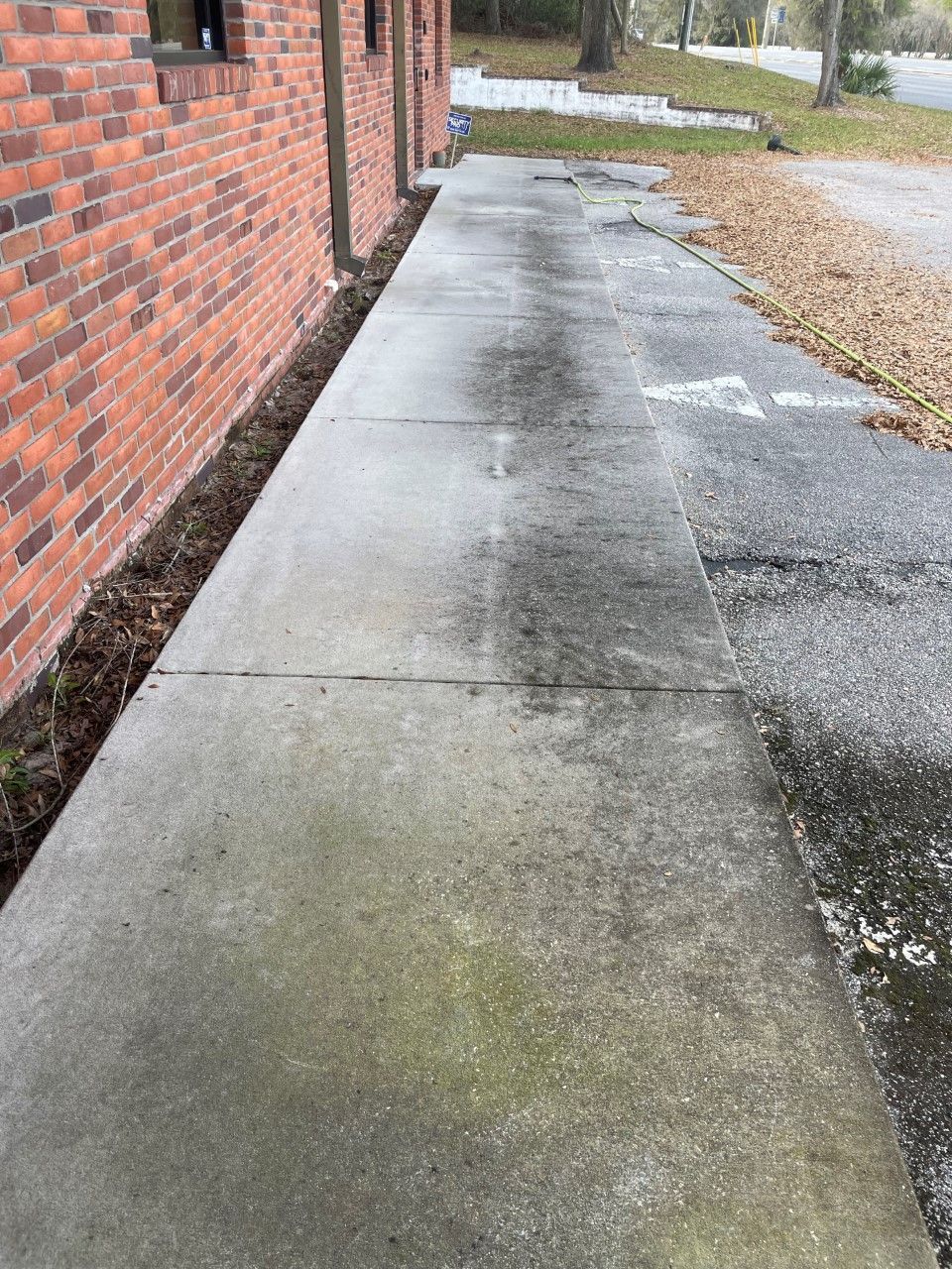 A concrete walkway runs along a brick building next to an asphalt parking area with a white painted directional arrow.