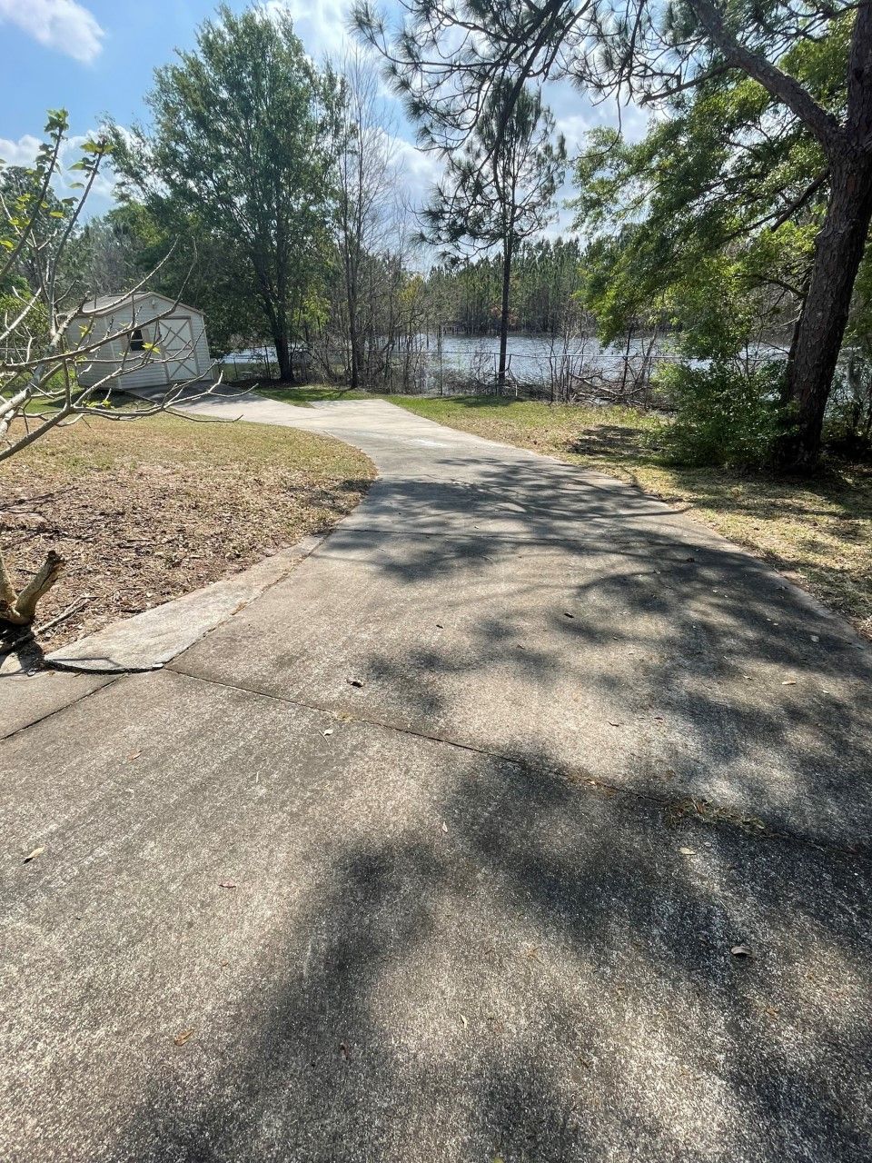 A concrete driveway leads toward a small shed and a body of water surrounded by trees under a blue sky.