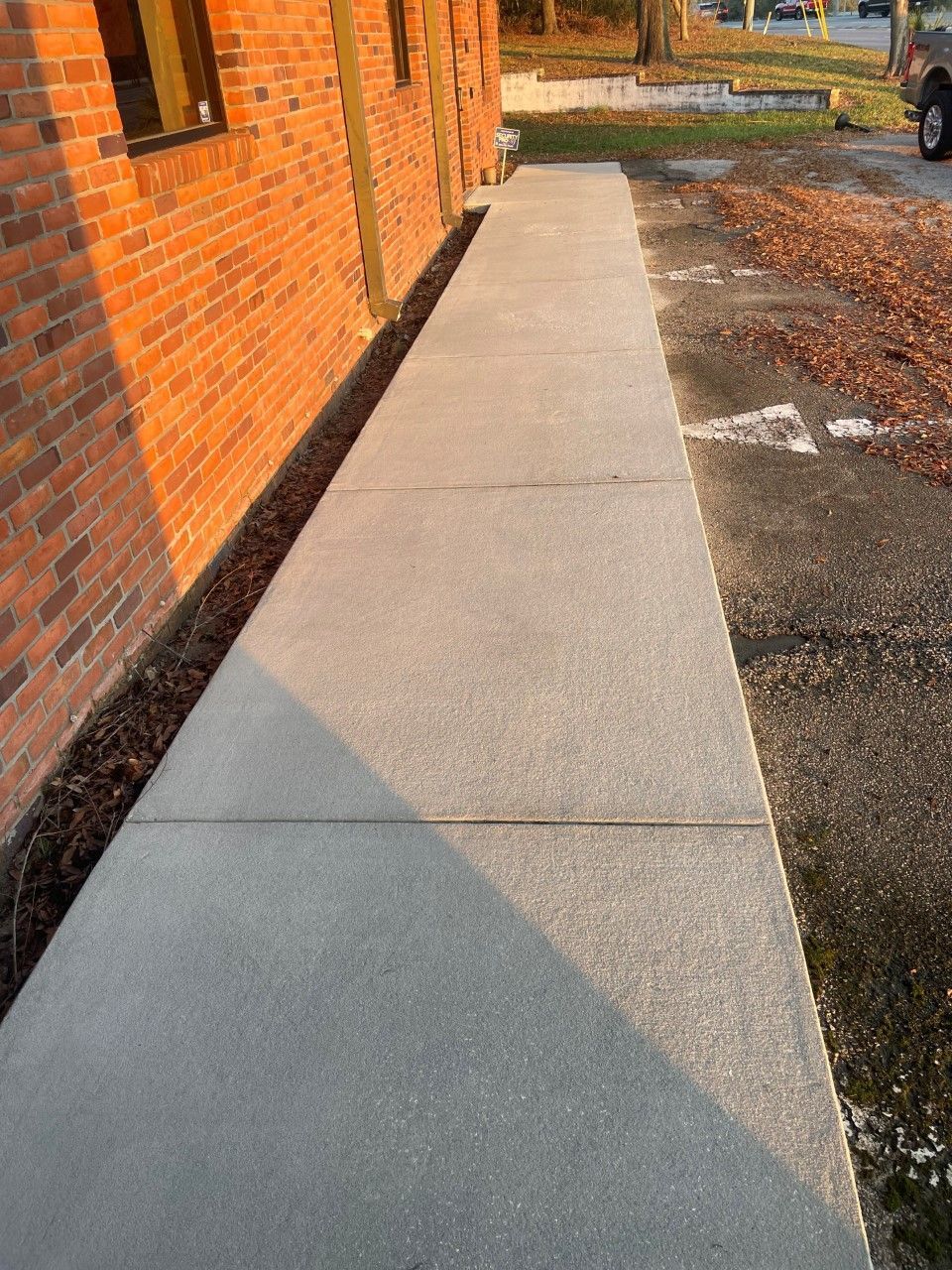 A newly poured gray concrete sidewalk runs alongside a brick building next to an asphalt parking lot.