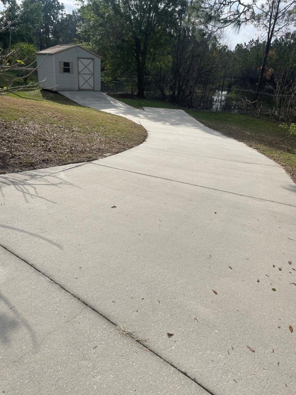 A paved concrete driveway leads up to a small, light-colored storage shed surrounded by trees and grass.