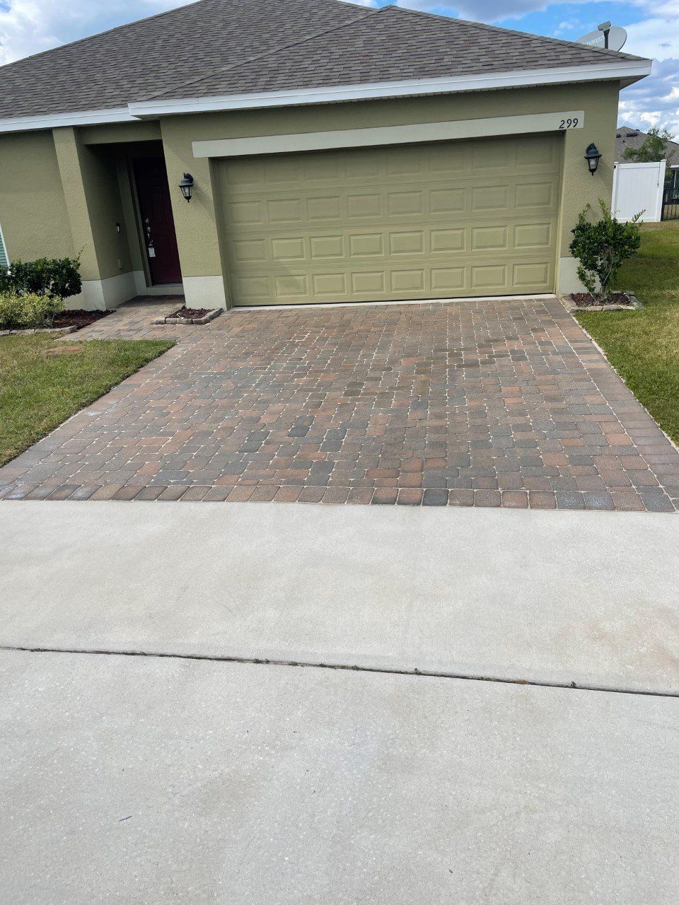 A single-story suburban house with a tan exterior, a brown shingled roof, and a paved stone driveway leading to a garage.