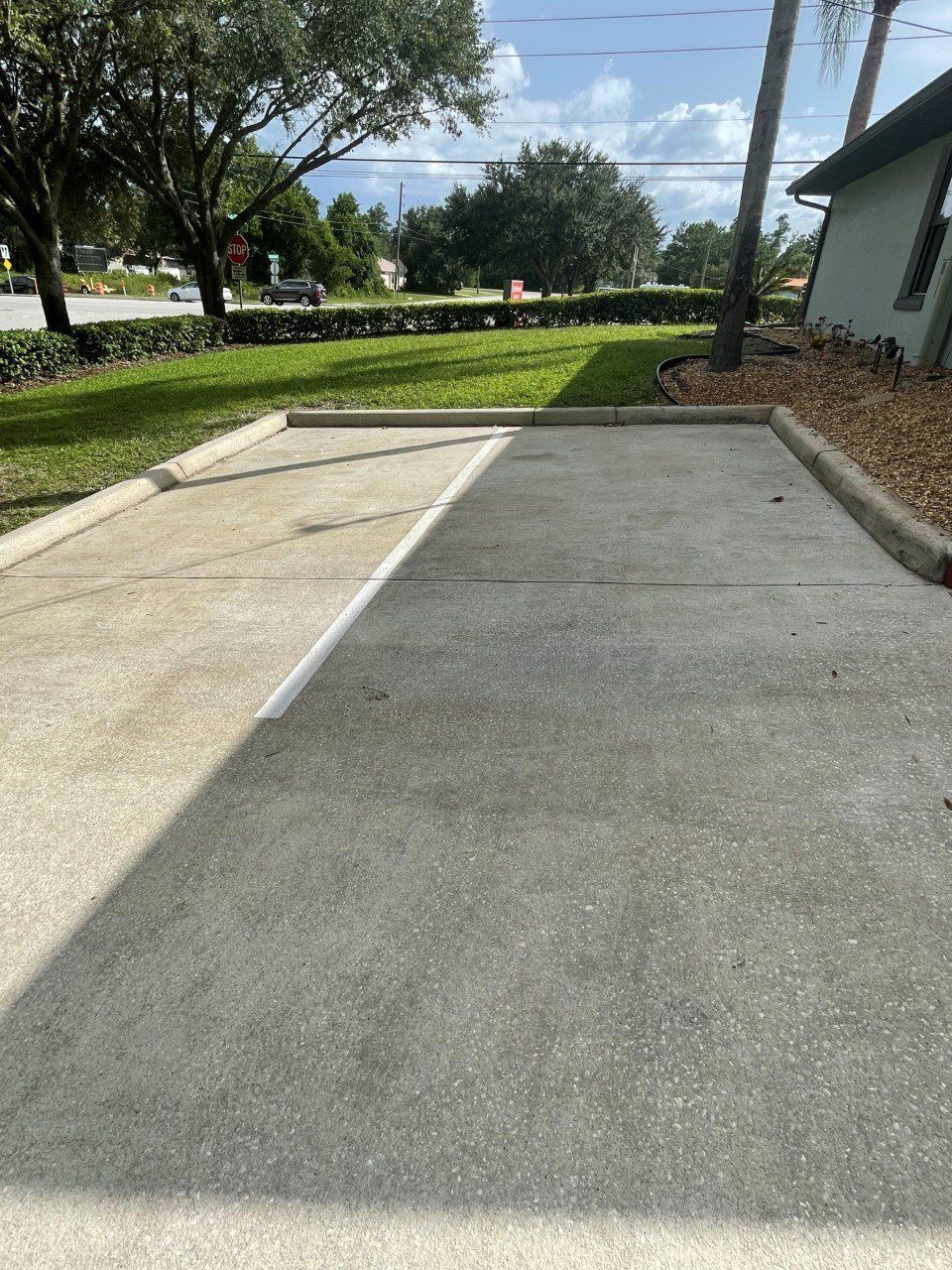 A parking space with concrete paving, divided by a shadow cast across the surface next to a grassy lawn.