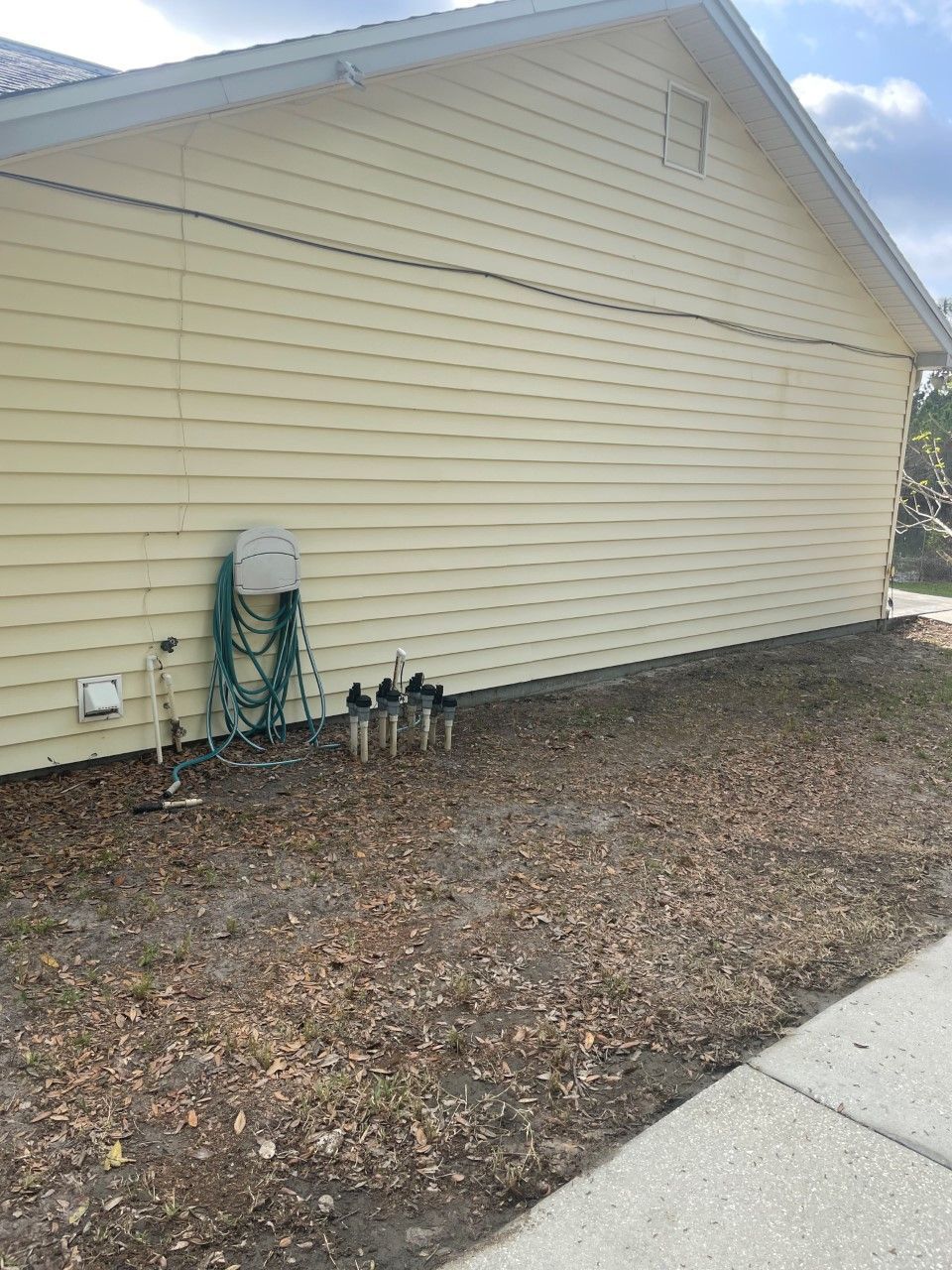 A light yellow vinyl-sided house exterior with a hose, spigot, and small utility pipes protruding from the ground.