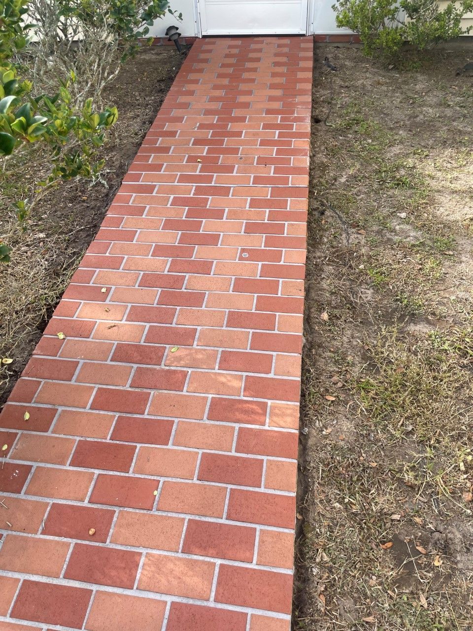 A straight outdoor walkway made of reddish-brown bricks, flanked by green bushes and dirt on both sides.