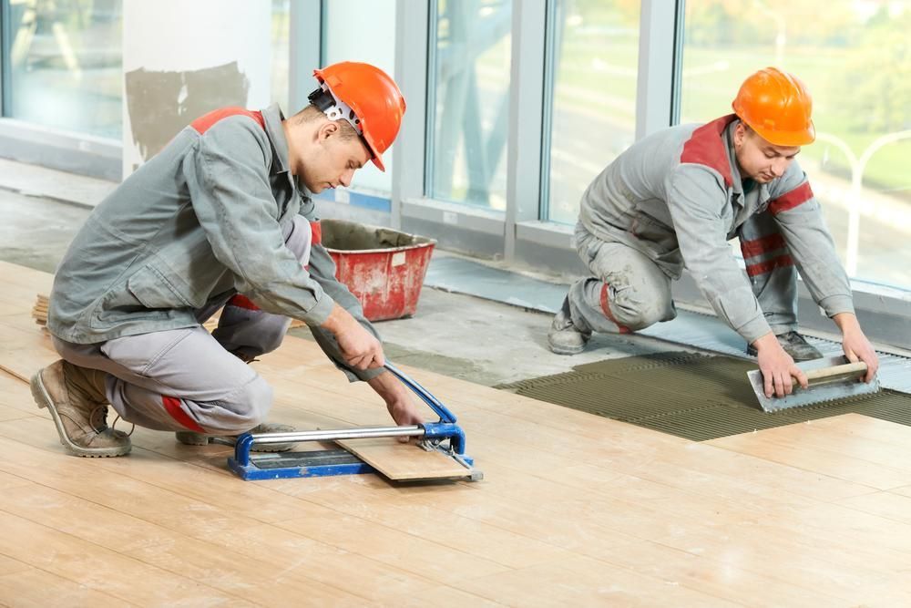 Two Men Are Installing Tiles On A Wooden Floor — B2B Flooring Solutions In Wauchope, NSW