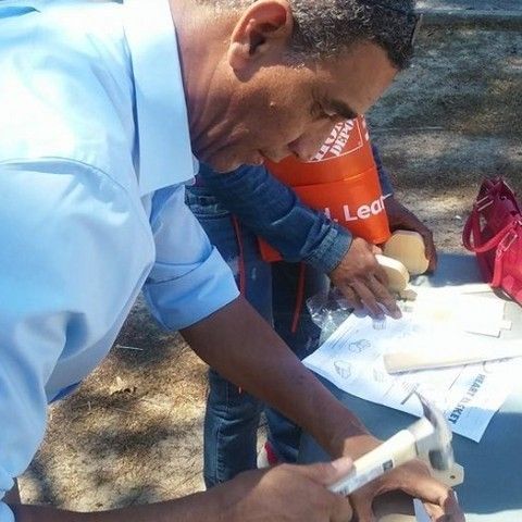 A person in a light blue shirt uses a hammer on a wooden piece at a table with a Home Depot bag and paper nearby.