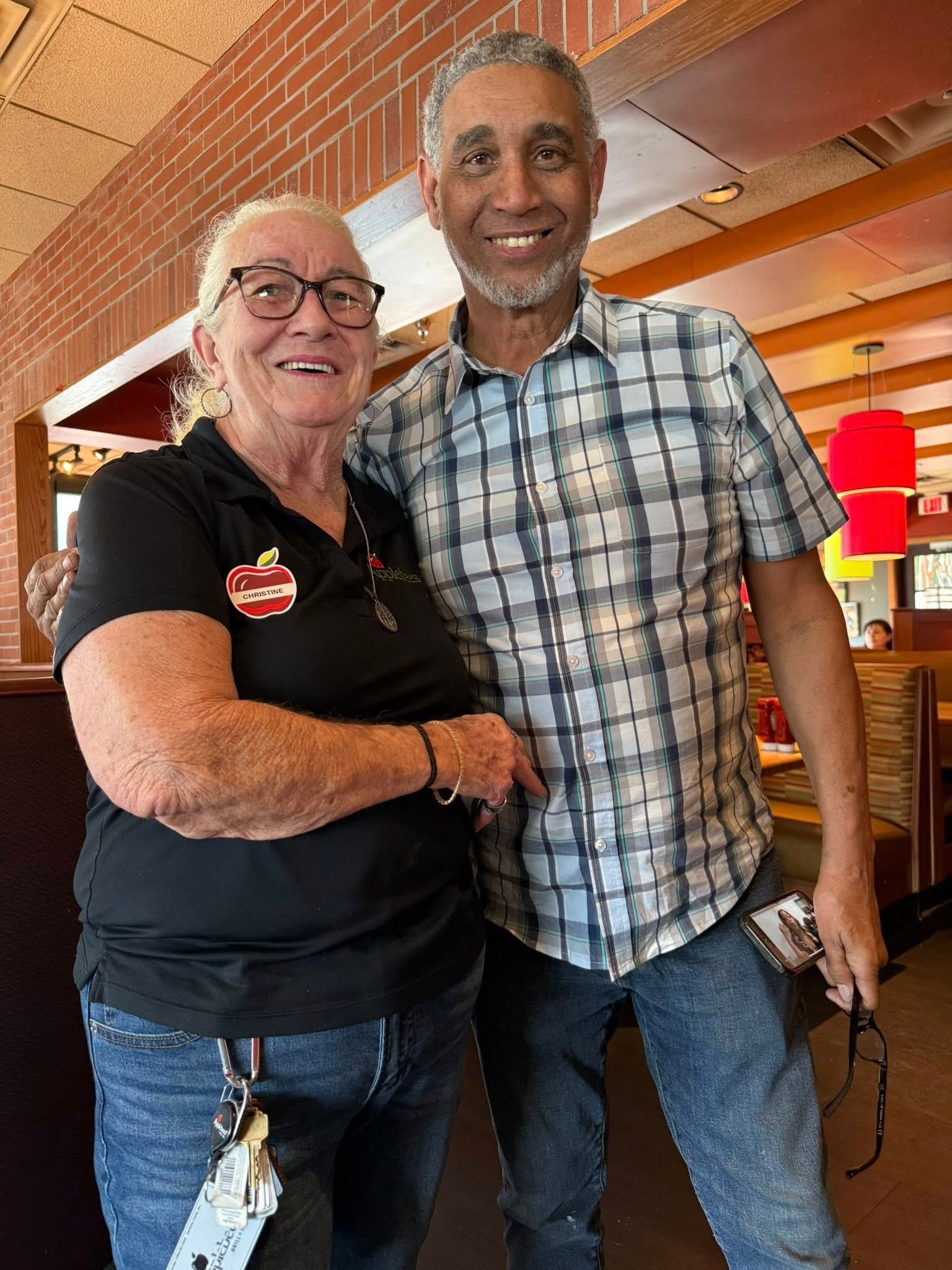 A smiling couple stands together indoors, wearing casual clothing, with the person on the left pointing to the other.