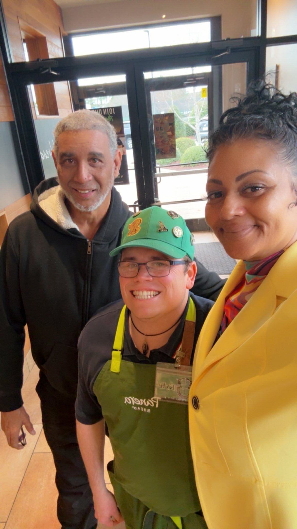 A selfie of three people smiling indoors. One person wears a green work apron and cap; others wear casual and formal jackets.