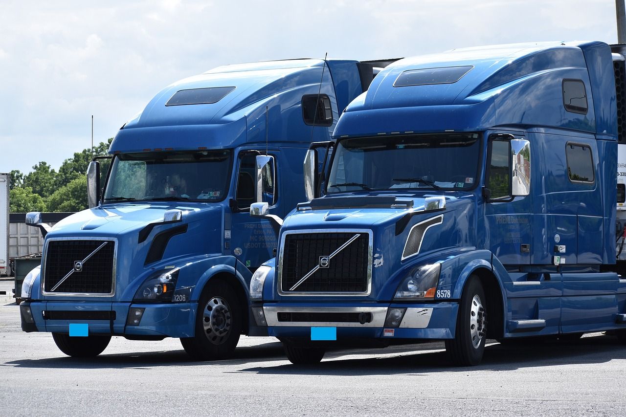Two blue semi-trucks parked side-by-side, with a bright sky in the background.