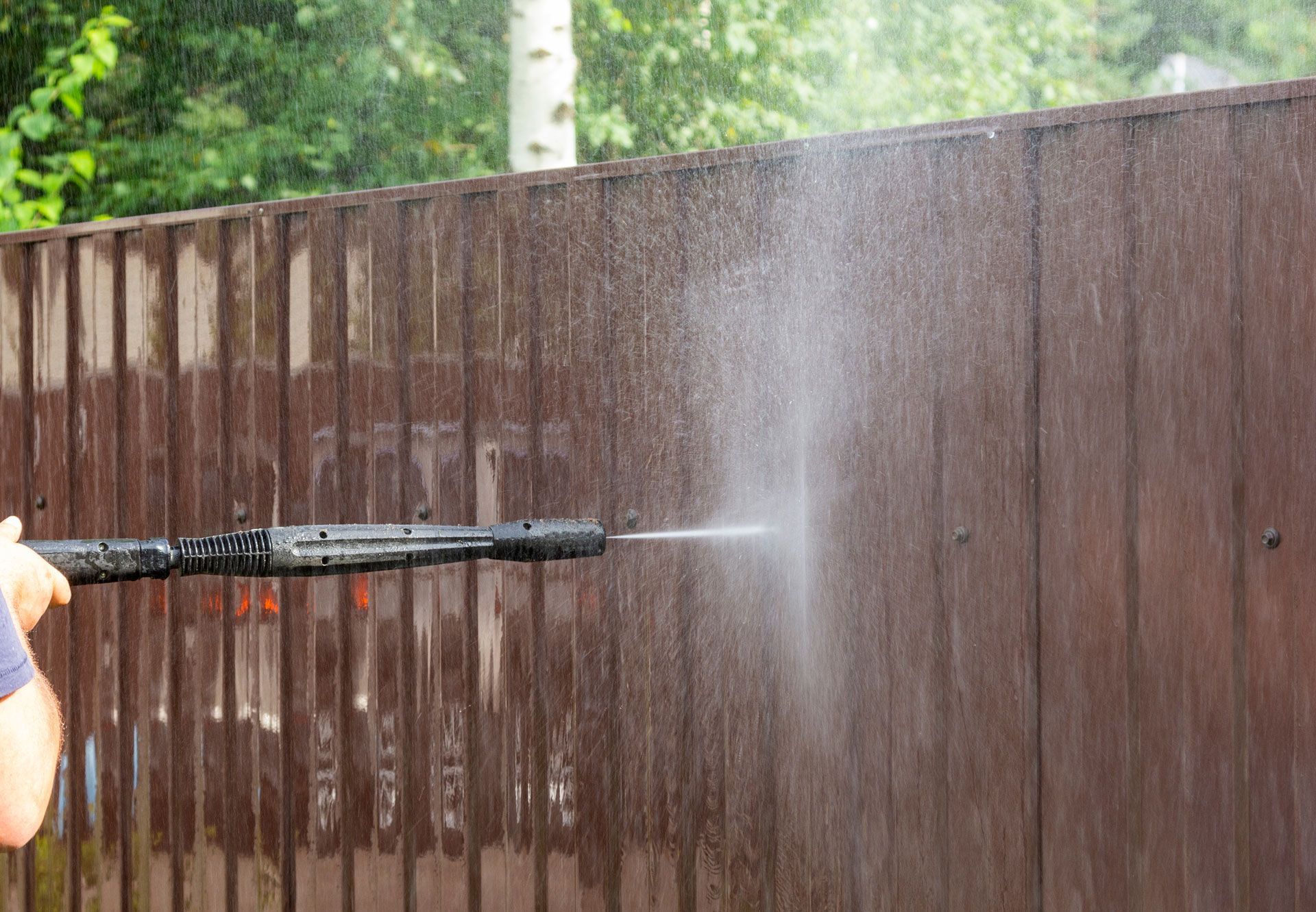Person power washing a brown metal fence with water spray.