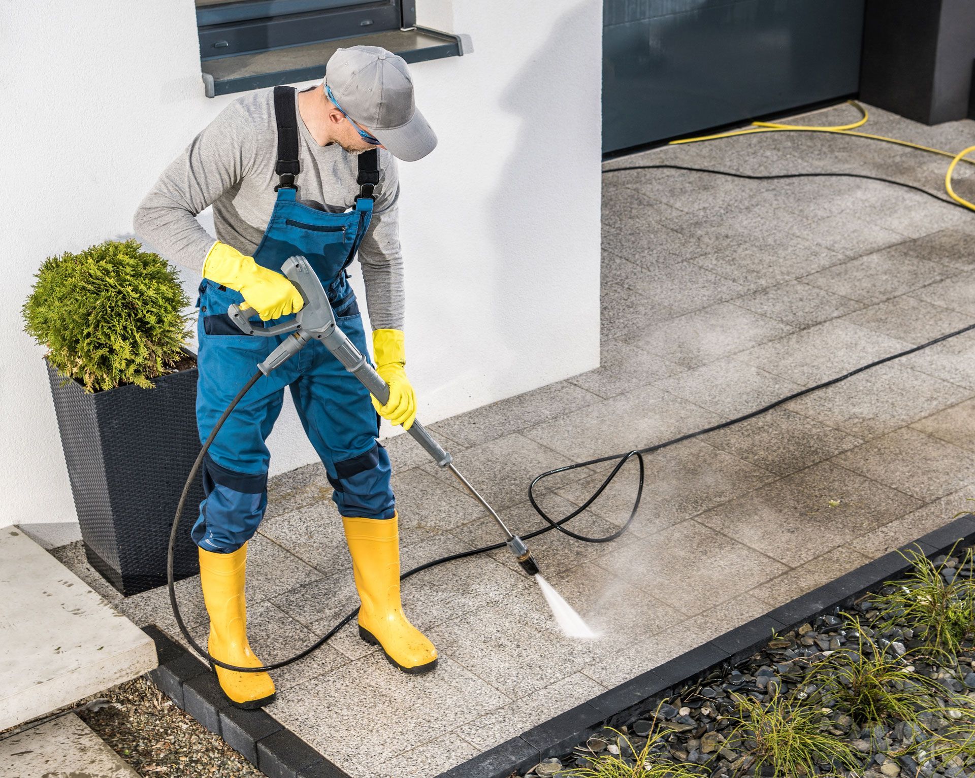 Person in yellow boots and gloves pressure washes a gray patio.