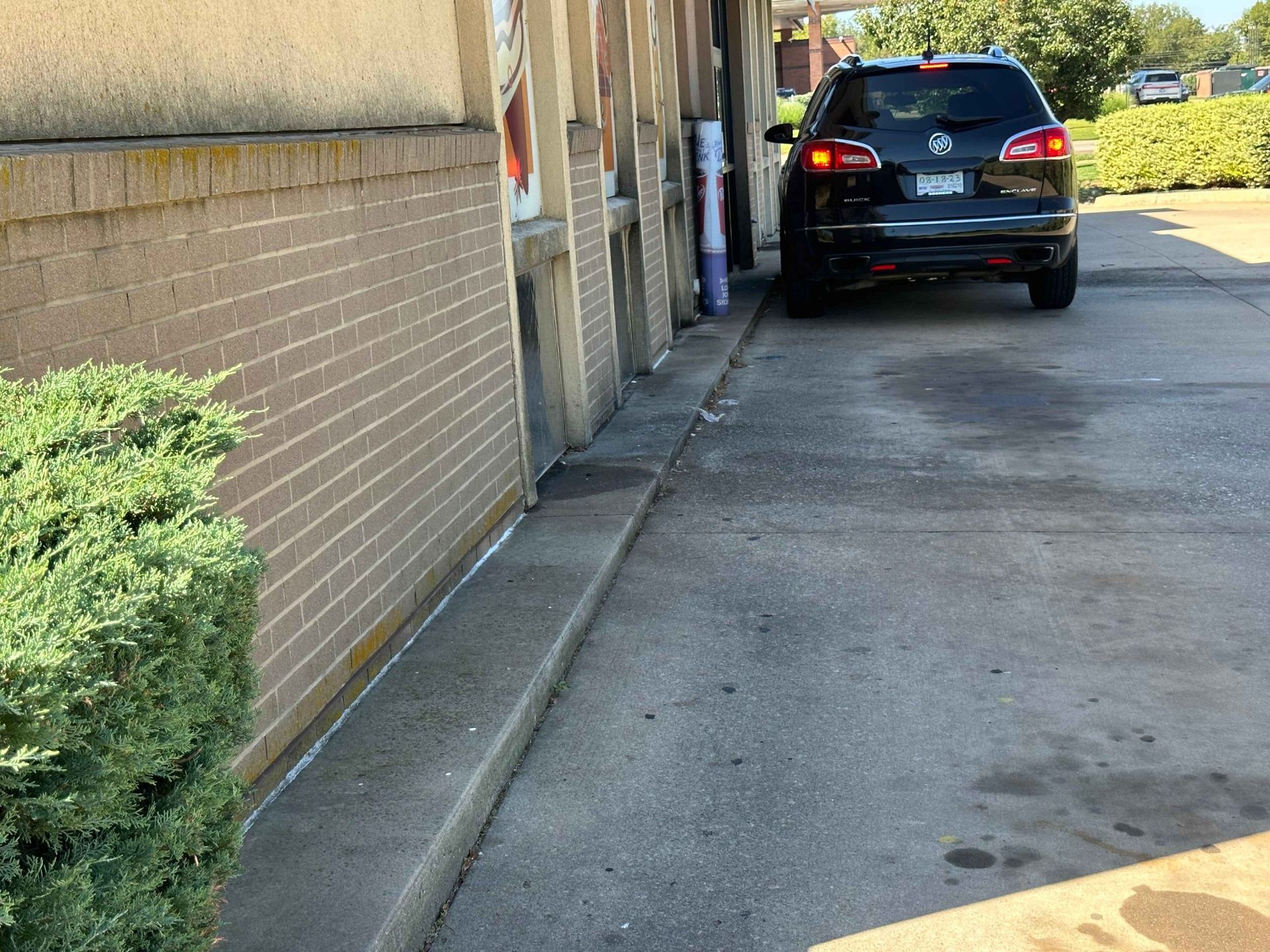 Black SUV parked next to a building, along a concrete walkway. Bush on the left.