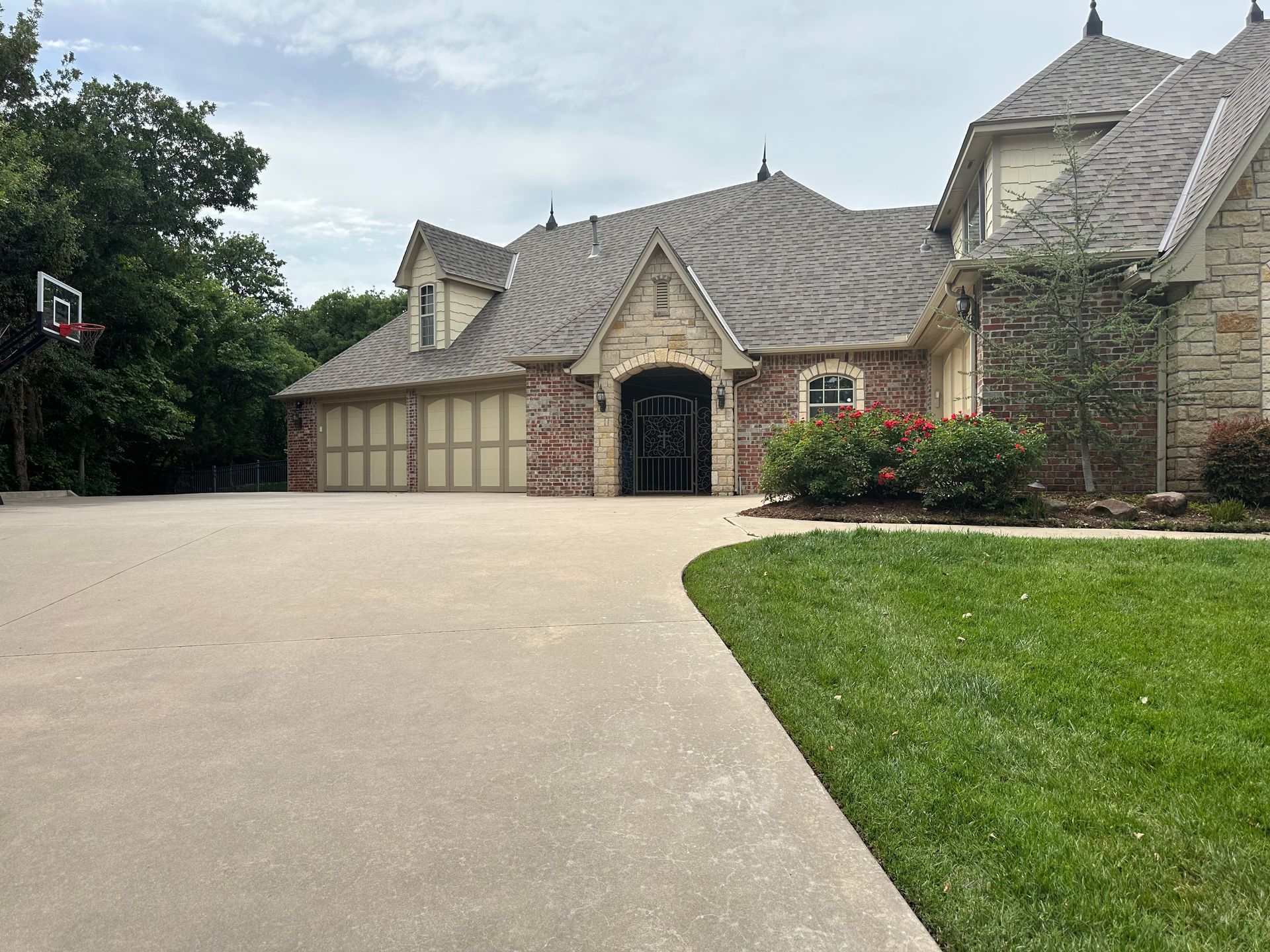 Large stone house with a steep gray roof, wide driveway, and green lawn under an overcast sky