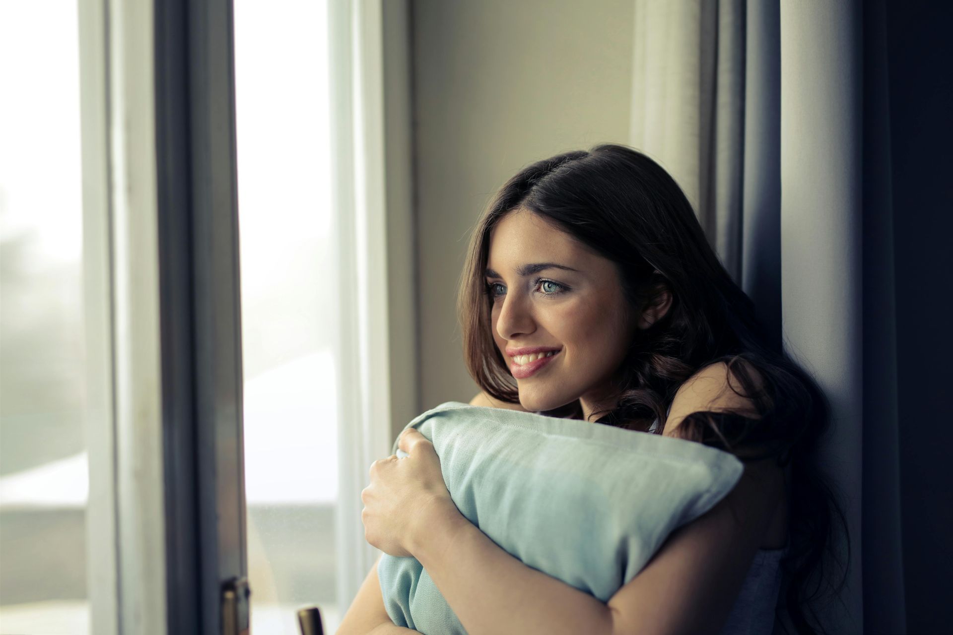A woman is holding a pillow and looking out of a window.