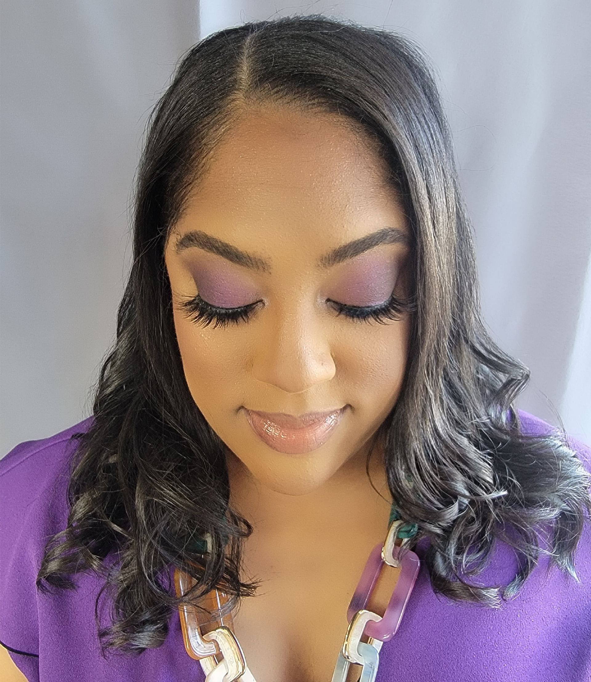 Woman with dark wavy hair and purple eyeshadow, looking down. Wearing a purple shirt and necklace.