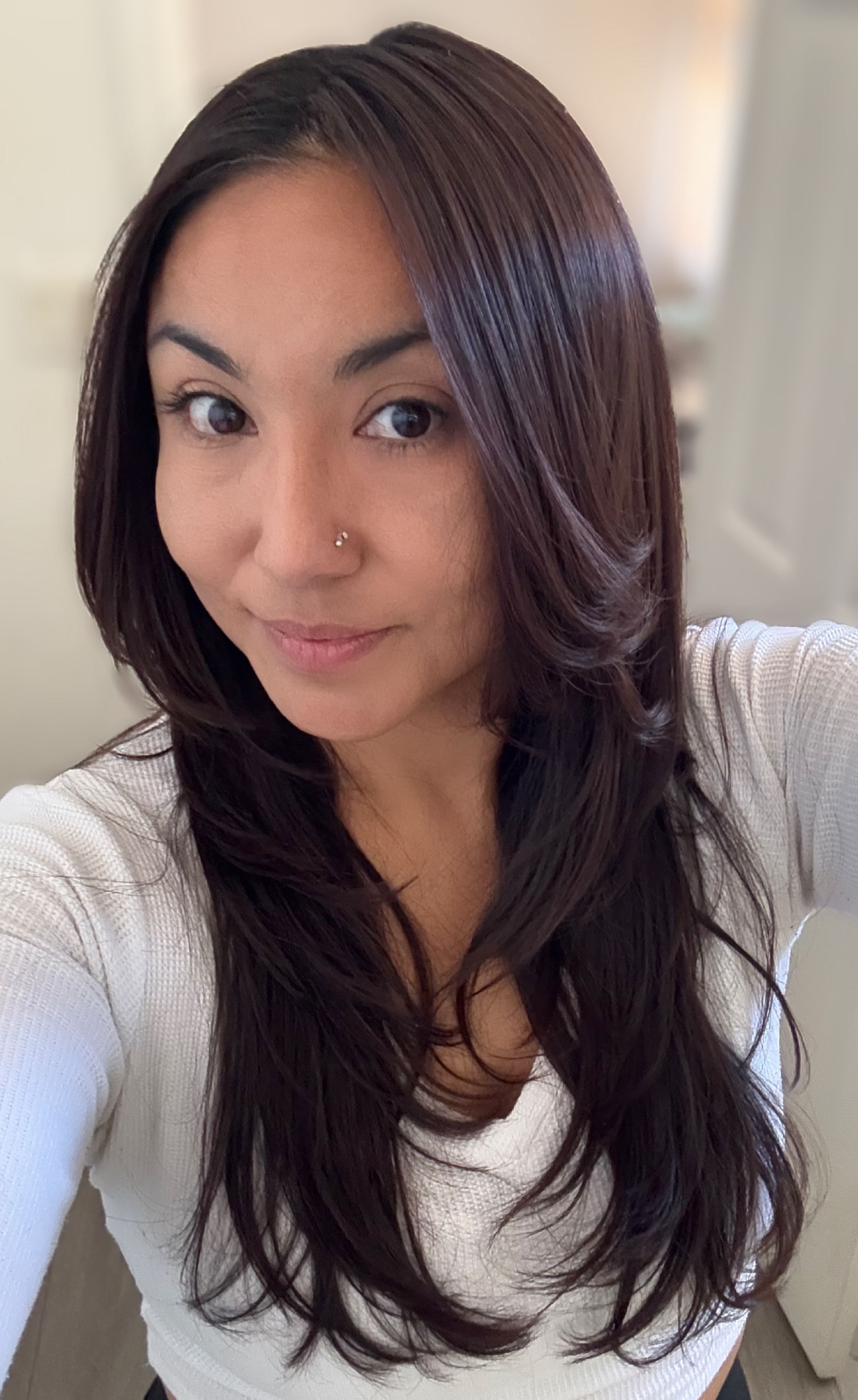Woman with long dark layered hair smiles, wearing a white top, indoors.