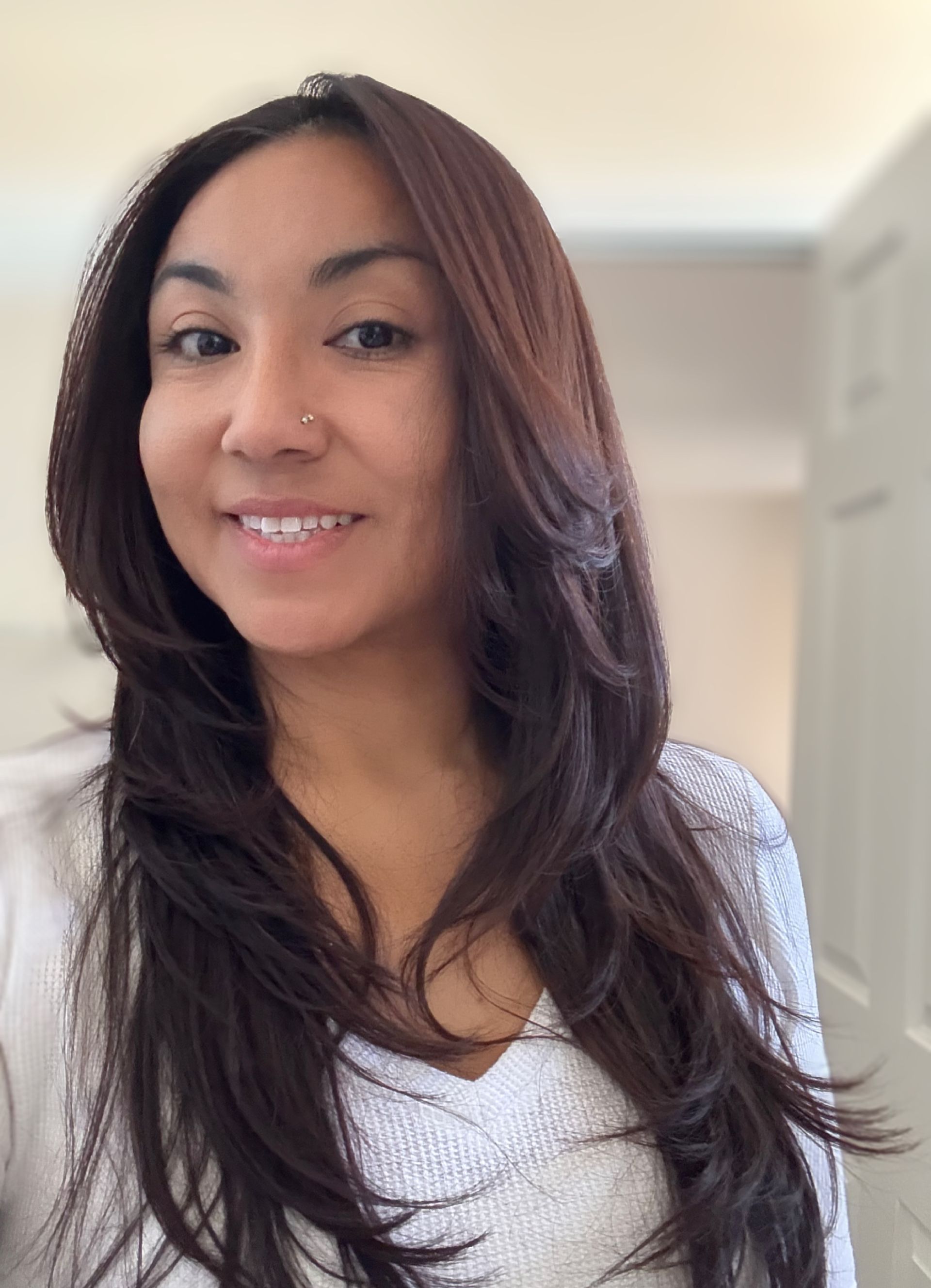 Woman with long, layered brown hair smiles. She wears a white sweater, and a nose ring. Indoors, blurred background.