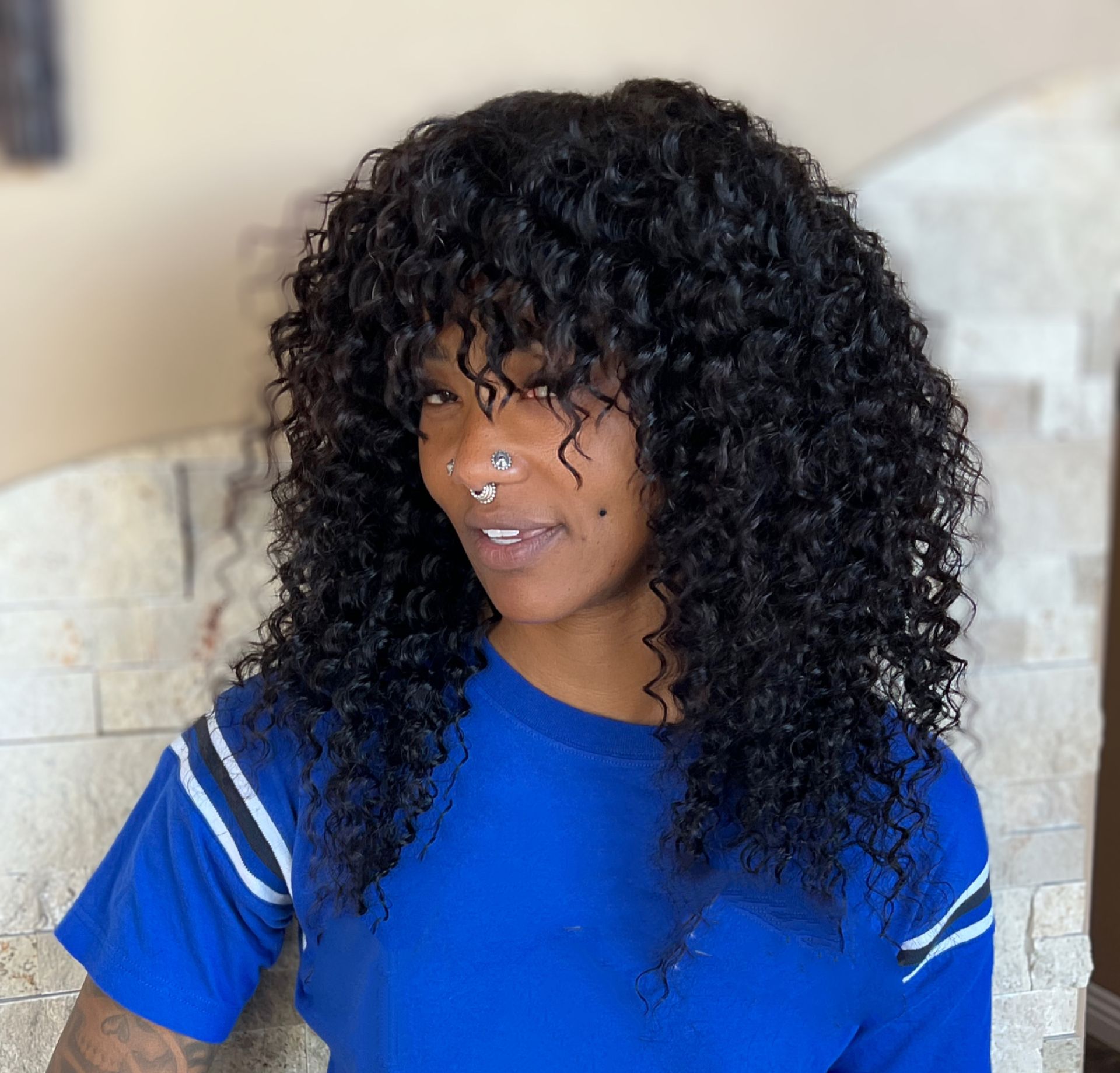 Person with curly black hair and bangs in a blue shirt smiling near a stone wall.