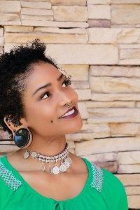 Woman with dark curly hair and jewelry looking up, wearing a green top, in front of a stone wall.