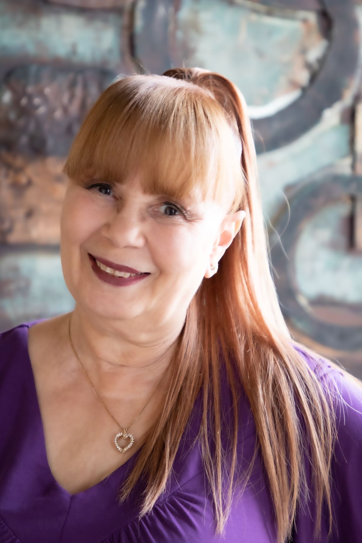 Woman with red hair and bangs, smiling, wearing a purple shirt, pendant necklace, in front of a textured wall.