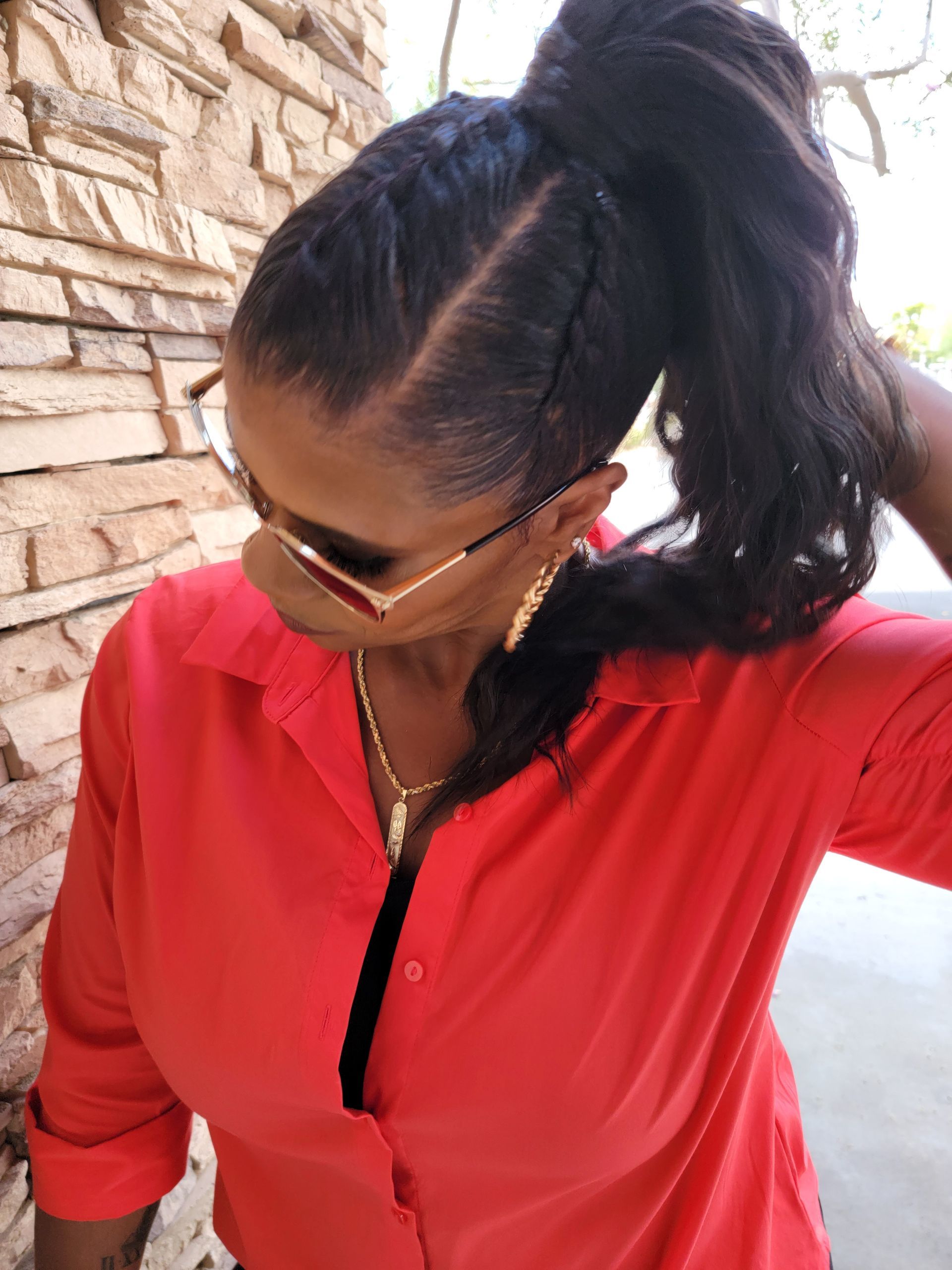 Woman with braided hair, wearing a red shirt, sunglasses, and a gold necklace, looking down.