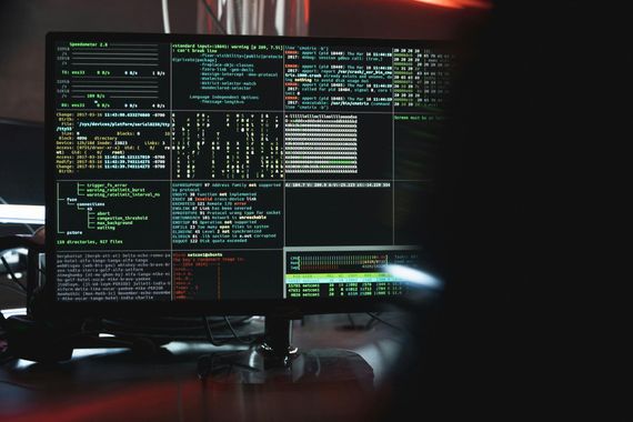 A computer monitor displays multiple terminal windows with code, command-line interfaces, and data logs in a dark room.