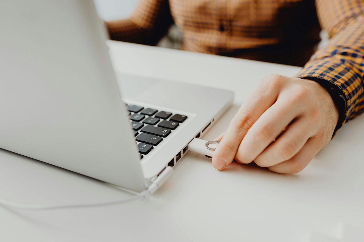 A person in a plaid shirt inserts a silver USB flash drive into the side of a laptop on a white desk.
