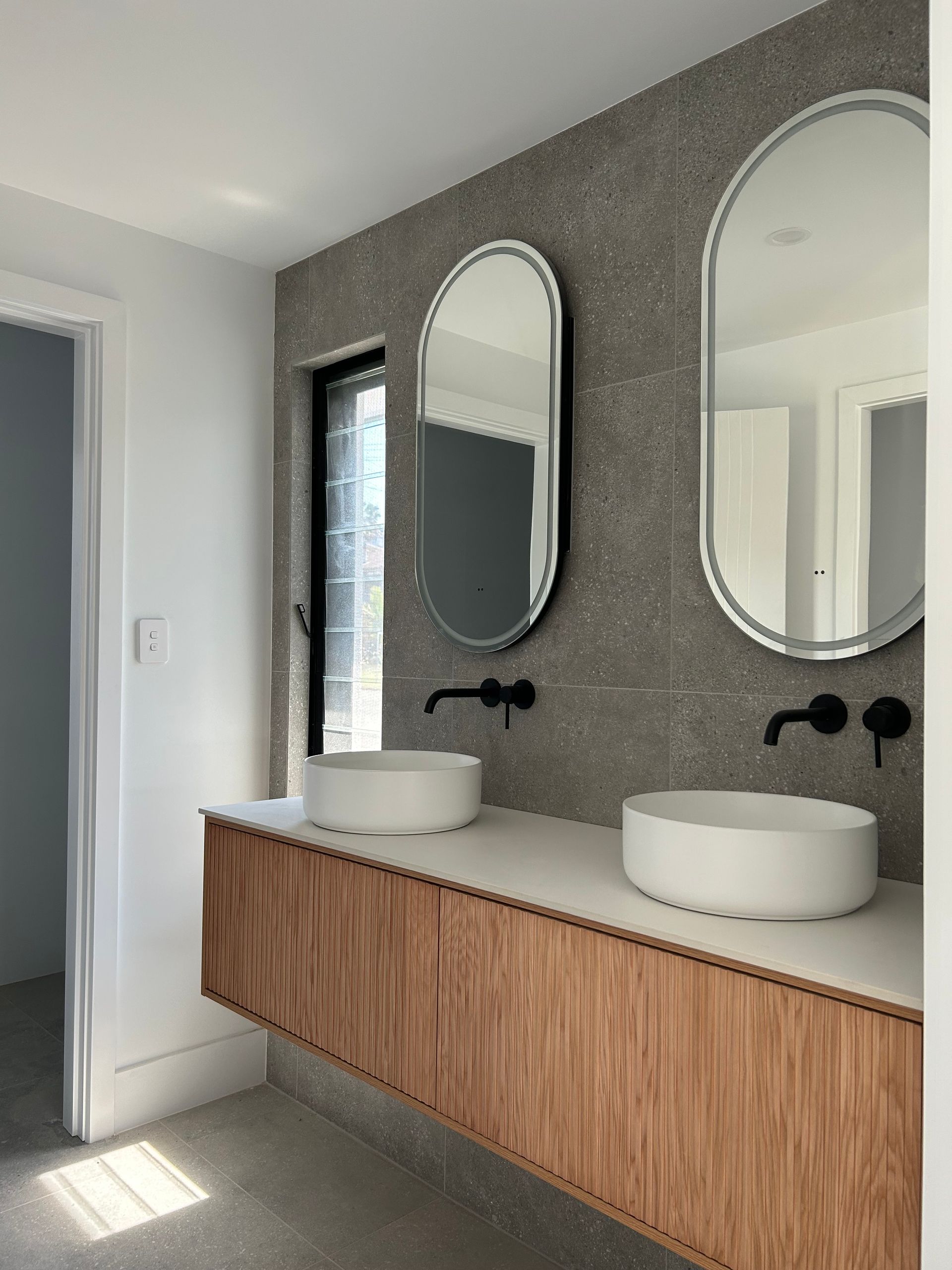 Modern bathroom with floating wood vanity, twin oval mirrors, and white vessel sinks on a grey wall — Wade Adams Constructions in Forster, NSW