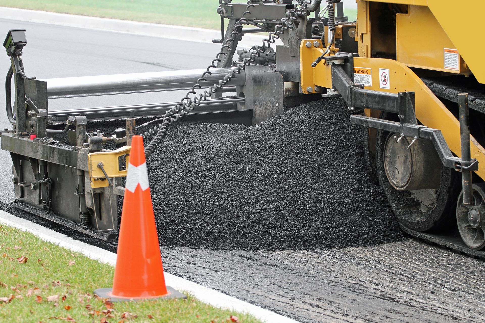 A construction machine is laying asphalt on a road next to an orange cone.
