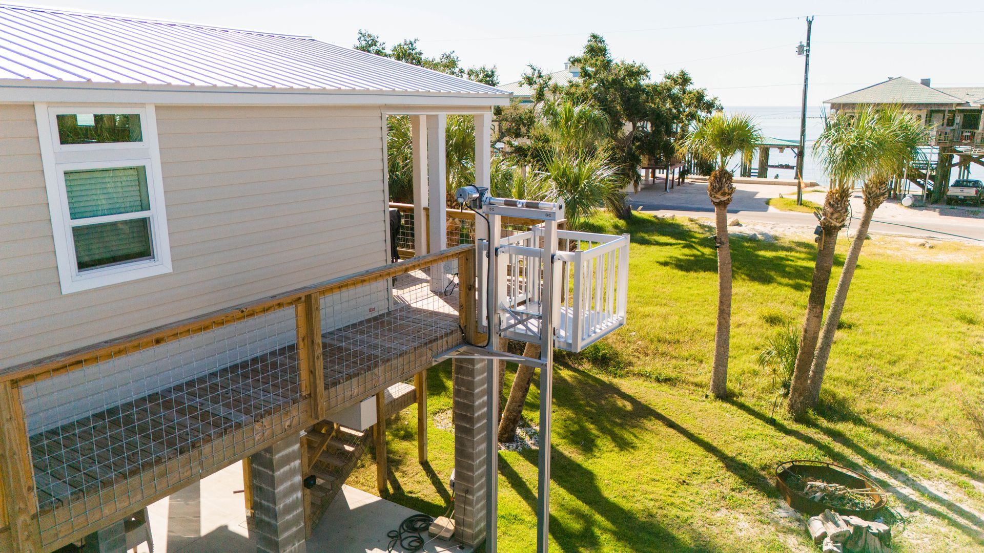 Beige beach house with elevated deck and ramp, near palm trees and water.