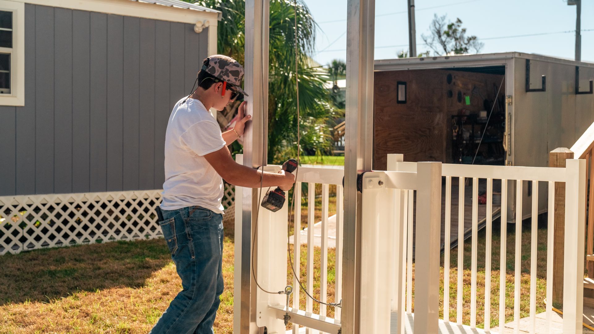 A person in a white shirt and jeans using a drill on a white picket fence in front of a mobile home.