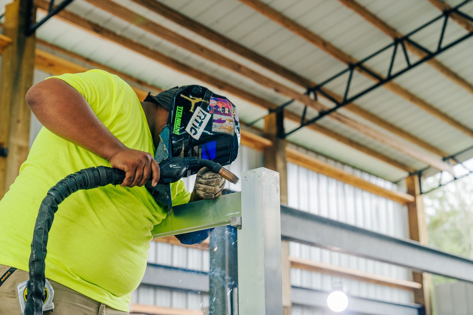 A construction worker welds metal while wearing a protective helmet and gear under a metal roof.