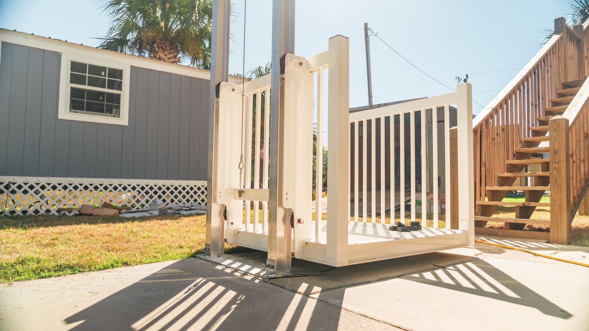 A white wheelchair lift outdoors, near a wooden staircase and a blue house. Sunny day.