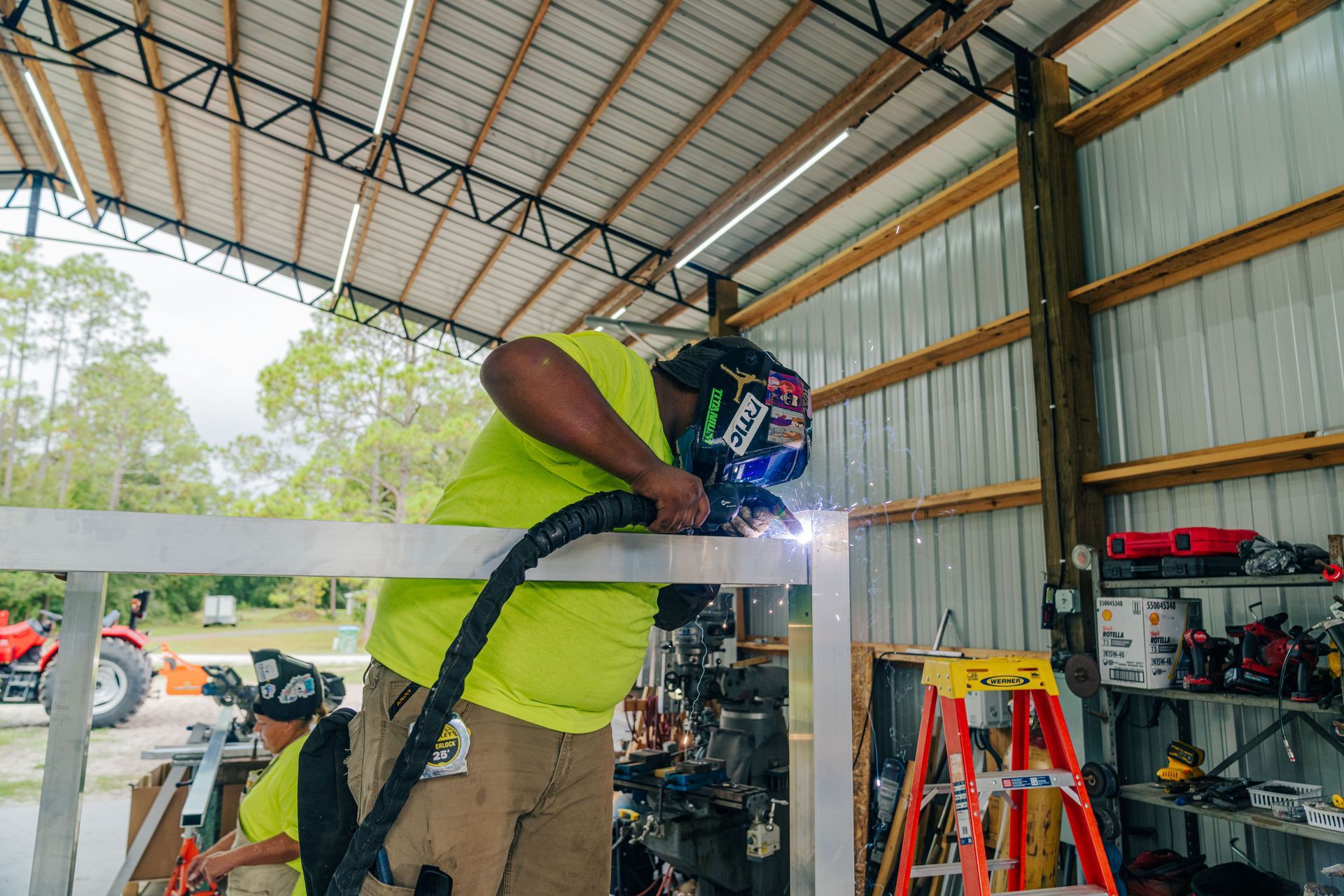 Two people welding metal in a workshop. One wears a yellow shirt and protective mask, the other in the background.