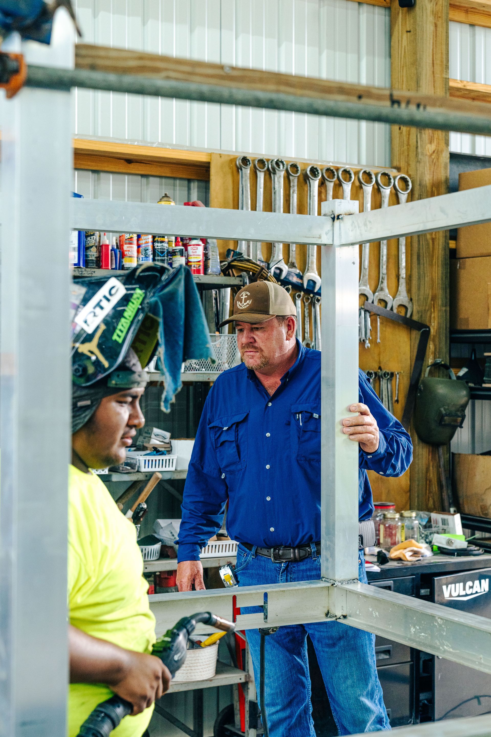 Two men in a workshop: one welding, the other observing a metal frame.