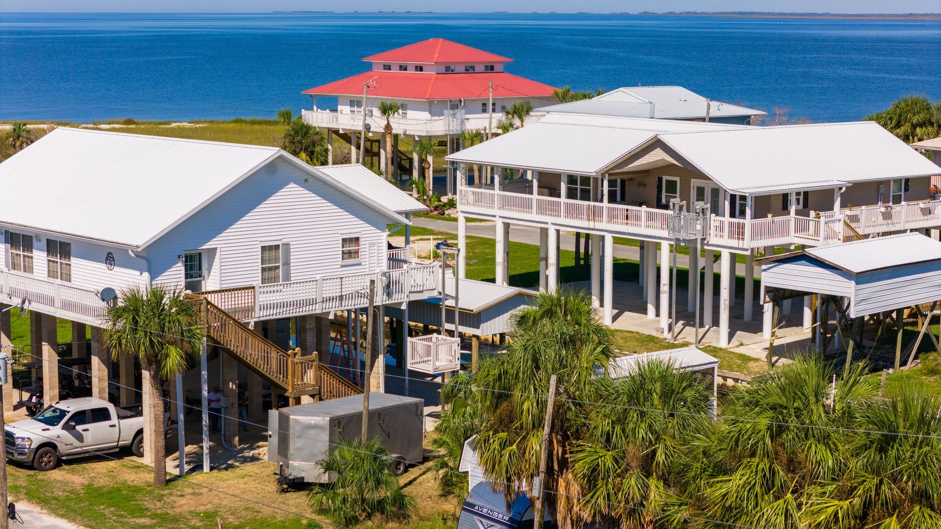 Beach houses on stilts with a red-roofed building in the background. Truck parked on the left.