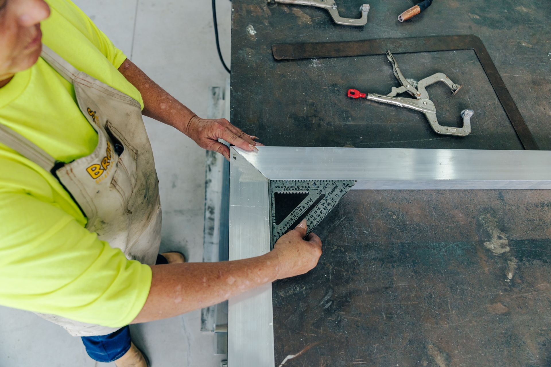 Person in yellow shirt using a square to measure metal frame on a workbench.